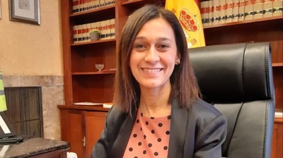 María Pilar Trinidad Núñez sentada en un despacho, con una bandera de España y estanterías de libros al fondo.